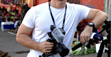 A young male journalist in a white tshirt with a camera around his neck leans against a tripod in a village street.